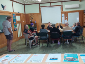 Ten people sitting around a table in a meeting room. There is a person standing to the left. In the foreground is a table with printed documents laid out.