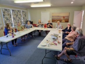 Nine people sit around a u-shaped table. There are plates, mugs and biscuits on the table. In the background are floral curtains and a beach scene painting.