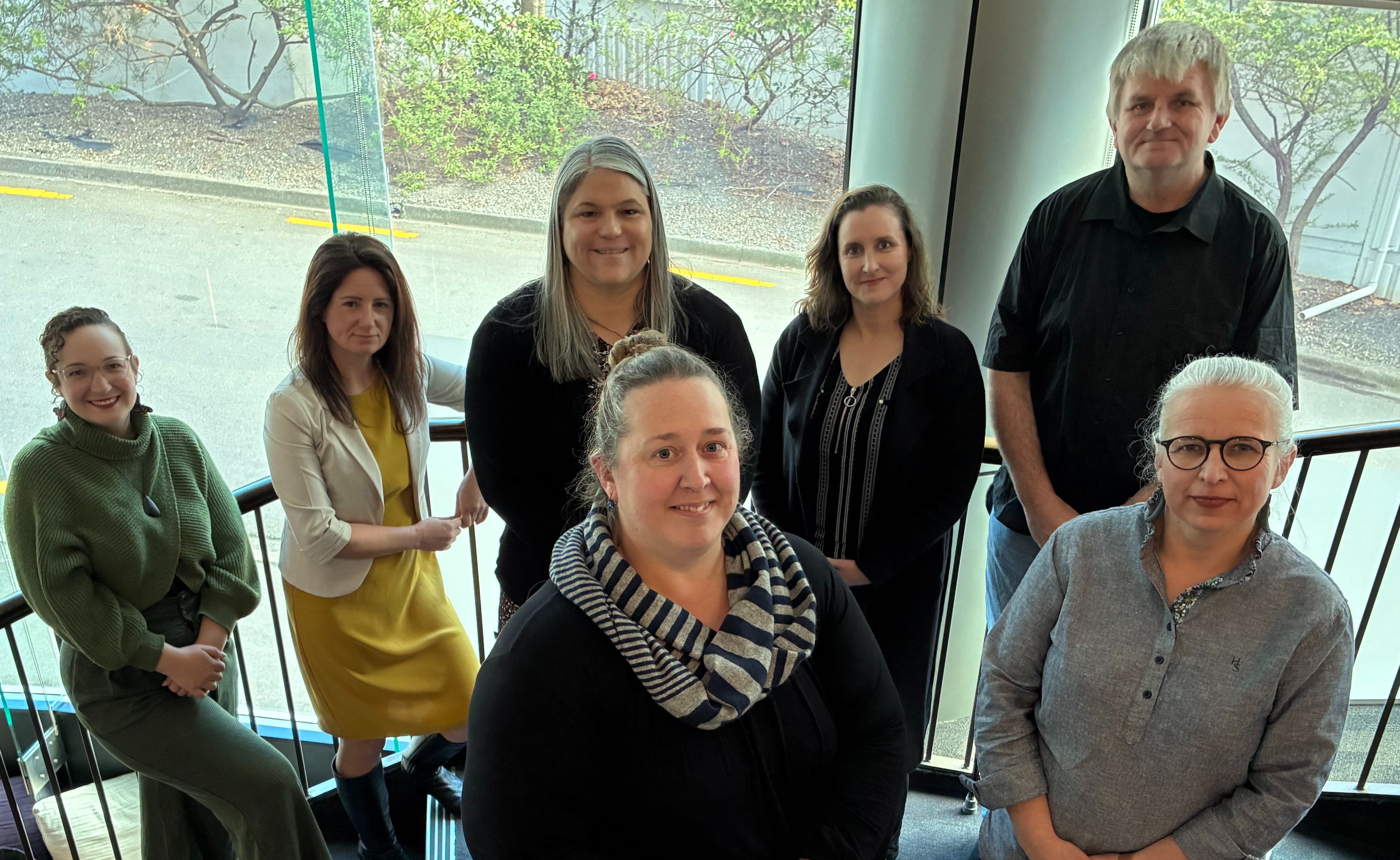 A group photo of the 7 NZSL Board members, standing in a stairwell. In the background is a road and trees.